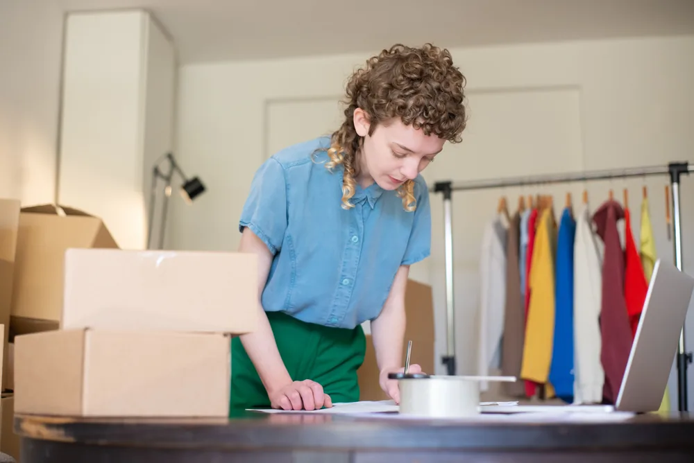 A woman writing on paperwork beside a laptop, clothing rail and packed boxes.